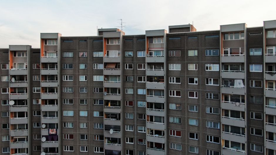 Exterior view of a multi-storey residential building in East Finchley with rows of balconies and windows, some with satellite dishes and laundry. In the foreground, the street level shows a sidewalk and a parking area. The building's facade is predominantly grey with small areas of orange accents around certain balcony sections. This setting relates to house removals and moving services in East Finchley, such as those provided by Man with Van East Finchley, which involves tasking furniture transportation and packing logistics for home relocations. The image captures a typical residential building that may be involved in local moving operations, emphasizing the context of apartment or flat relocations along High Road flats N2 in East Finchley.