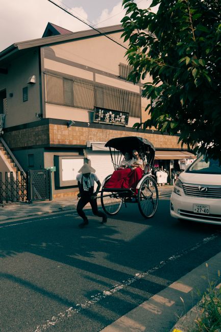 A traditional rickshaw with a large red seat cover is being pulled along a city street by a person wearing a conical hat and a light-colored shirt. Inside the rickshaw, a passenger is seated under a black canopy, carrying a bag and appearing to be resting or waiting. The street features parked cars, including a silver sedan on the right, and a modern multi-storey building with balconies and a tiled exterior in the background. The pavement has white markings, and a tree with green leaves partially obscures the upper right of the image. This scene captures a typical urban area, illustrating transportation methods and moving preparations, which May be part of packing or furniture transport related to home relocation services by Man with Van East Finchley in the context of house removals.