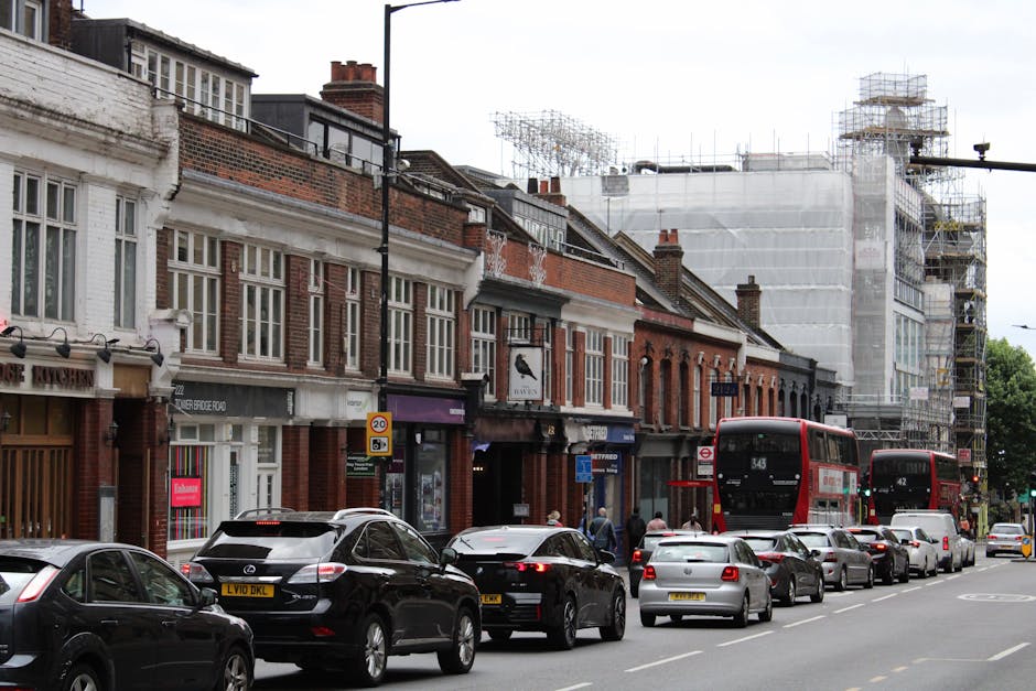 Exterior view of a multi-storey residential building in East Finchley with rows of balconies and windows, some with satellite dishes and laundry. In the foreground, the street level shows a sidewalk and a parking area. The building's facade is predominantly grey with small areas of orange accents around certain balcony sections. This setting relates to house removals and moving services in East Finchley, such as those provided by Man with Van East Finchley, which involves tasking furniture transportation and packing logistics for home relocations. The image captures a typical residential building that may be involved in local moving operations, emphasizing the context of apartment or flat relocations along High Road flats N2 in East Finchley.