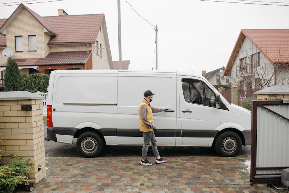 A man wearing a black cap, glasses, a yellow and grey jacket, and grey trousers is standing outside a residential property in East Finchley, on a paved driveway made of multicoloured bricks. He is opening the sliding side door of a white commercial van used for home relocation or furniture transport, which is parked parallel to the house. The van's rear and side doors are closed, and the vehicle is positioned in front of a low brick wall with a garden area visible beyond it. Nearby, a metal gate partially opening onto the driveway is closed, and the street features other detached houses with pitched roofs, some with dormer windows, and suburban greenery. The scene suggests a loading process for moving furniture or household items, with the van prepared for packing or furniture loading, supported by the presence of packing materials and furniture blankets potentially used during the move, in the context of professional removals services by Man with Van East Finchley.