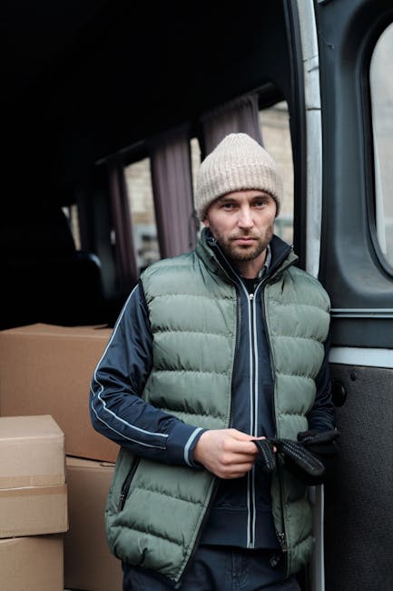 A man with a neutral expression, wearing a grey knit beanie and a green and navy puffer vest over a dark jacket, standing inside the rear of a moving van parked outside a residential property. Behind him, there are cardboard boxes stacked on the vehicle's floor, indicating a home relocation or packing process. The van's interior features dark upholstery and a window with a curtain, suggesting it is being used for furniture transport and packing during a house removal. The man appears to be preparing items for loading or unloading, with a gloved hand holding a strap or rope used to secure or carry belongings. Natural daylight illuminates the scene, emphasizing the professional handling of moving tasks by [COMPANY_NAME], which offers comprehensive removal and moving services in East Finchley, related to house removals, furniture transport, and packing and moving logistics.