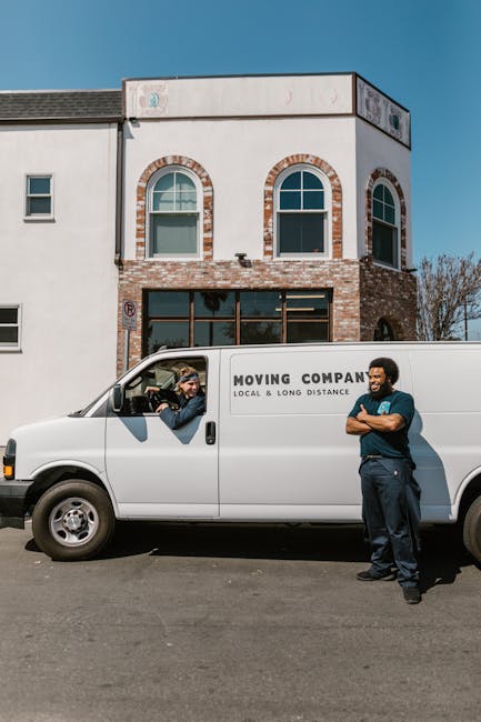 A white moving van with the branding 'MOVING COMPANY LOCAL & LONG DISTANCE' parked in front of a two-story residential building with a brick and white exterior. The building features arched windows on