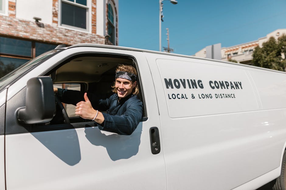 A white moving van with the branding 'MOVING COMPANY LOCAL & LONG DISTANCE' parked in front of a two-story residential building with a brick and white exterior. The building features arched windows on